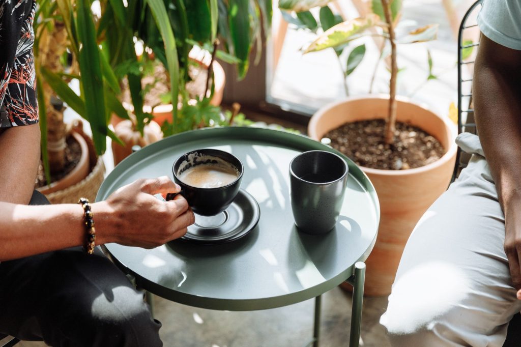 Deux personnes discutent autour d'une table basse avec deux tasses de café dans un espace lumineux agrémenté de plantes en pot.