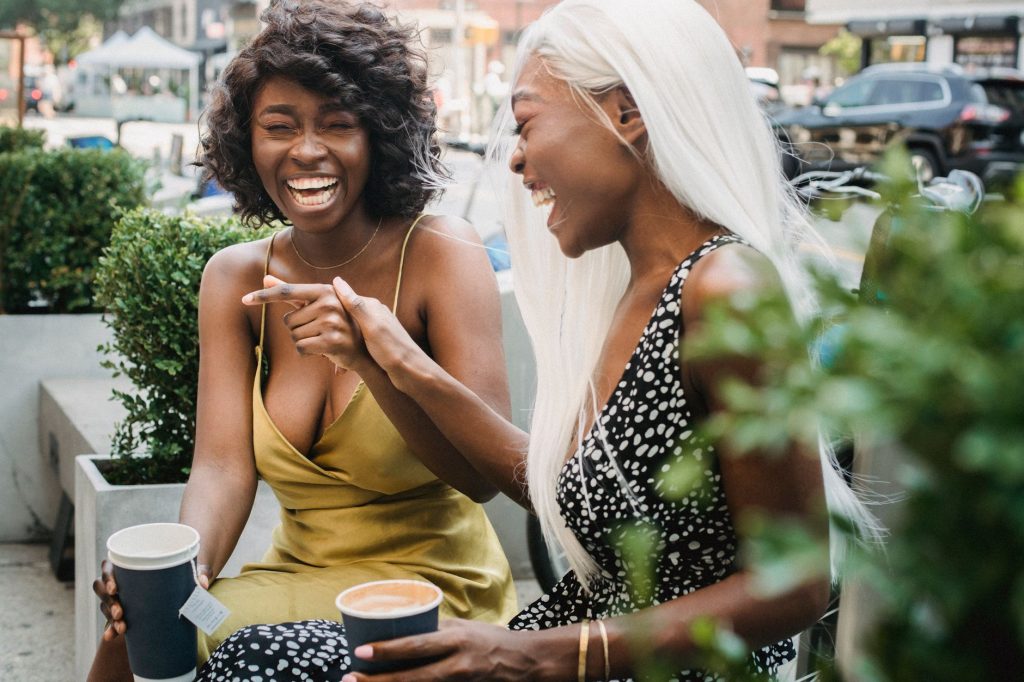 Deux amies riant ensemble en tenant des tasses de café à une table en terrasse.