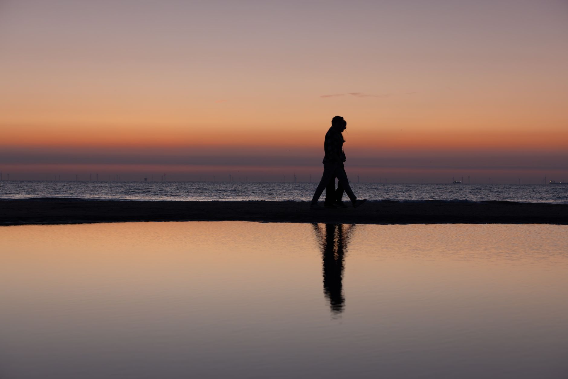 Silhouette d'un couple marchant main dans la main sur une plage au coucher du soleil avec leur reflet dans l'eau.